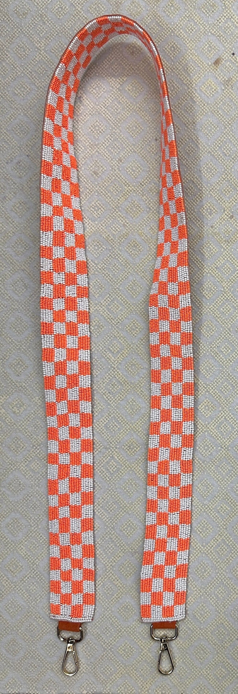May include: A long orange and white checkered strap with gold metal clasps. The strap is made of woven beads.