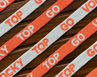 May include: Orange and white beaded ribbon with the text "Rocky Top Go Vols" repeated on it. The ribbon is on a brown wooden surface.