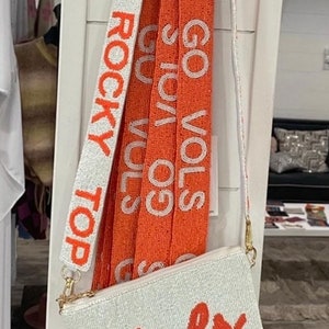 May include: A white beaded purse with an orange "vols" logo. The purse has a long beaded strap with the words "Top Rocky Top" and "Go Vols" in orange.