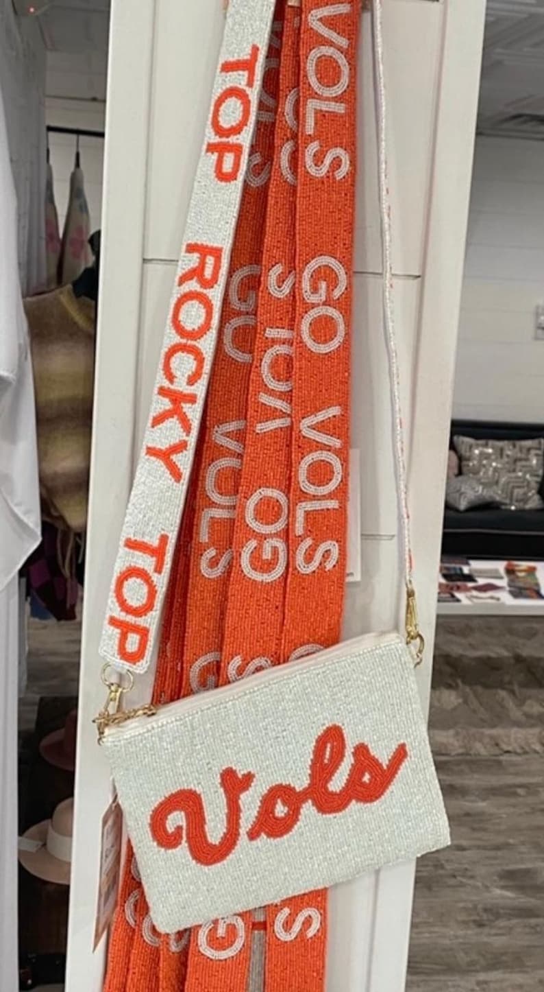 May include: A white beaded purse with an orange "vols" logo. The purse has a long beaded strap with the words "Top Rocky Top" and "Go Vols" in orange.