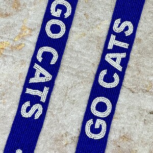 K&ouml;nnte beinhalten: Zwei blaue Perlenb&auml;nder mit wei&szlig;en Perlen, die "GO CATS" buchstabieren und wei&szlig;e Pfotenabdr&uuml;cke aufweisen.