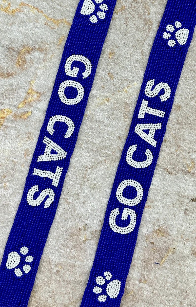 K&ouml;nnte beinhalten: Zwei blaue Perlenb&auml;nder mit wei&szlig;en Perlen, die "GO CATS" buchstabieren und wei&szlig;e Pfotenabdr&uuml;cke aufweisen.