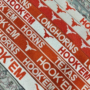 May include: A close-up of a stack of orange and white beaded belts with the words "Longhorns", "Hook 'Em", and "Texas" repeated on the belts. The belts are decorated with a stylized Longhorn steer head.