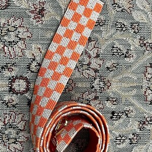 May include: A white and orange checkered beaded bag strap with a gold clasp.