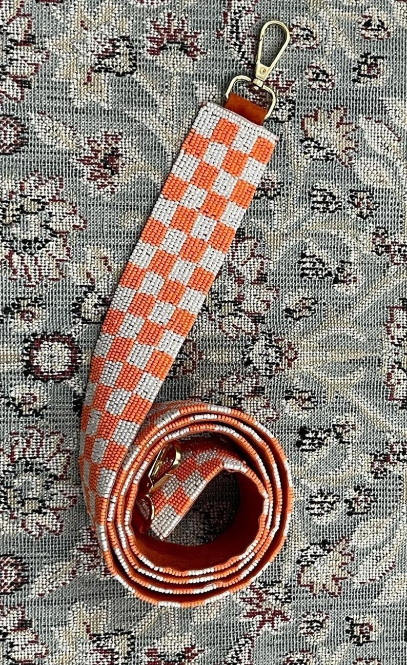 May include: A white and orange checkered beaded bag strap with a gold clasp.