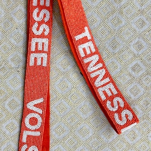 May include: Orange beaded bag strap with white lettering that reads "VOLS TENNESSEE". The strap has two gold clasps.