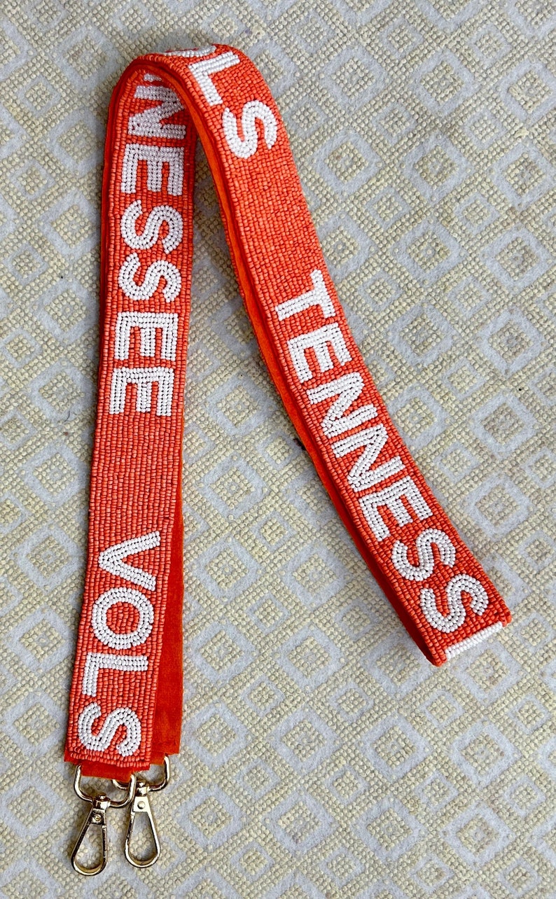May include: Orange beaded bag strap with white lettering that reads "VOLS TENNESSEE". The strap has two gold clasps.