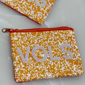 May include: A small orange zippered pouch with a gold zipper. The pouch is covered in gold and white sequins and has the word "VOLS" spelled out in white sequins.