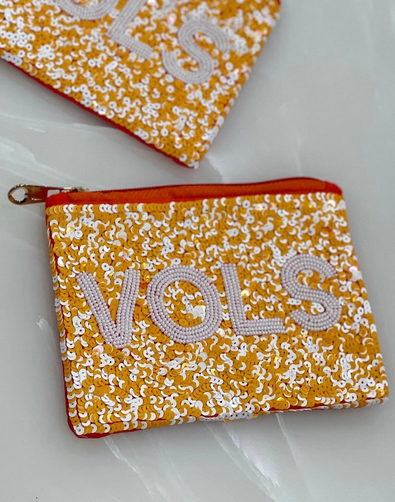 May include: A small orange zippered pouch with a gold zipper. The pouch is covered in gold and white sequins and has the word "VOLS" spelled out in white sequins.