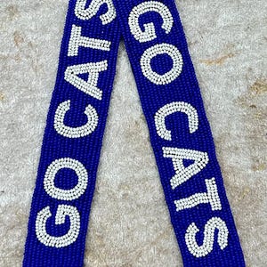 K&ouml;nnte beinhalten: Ein blaues Perlenband mit wei&szlig;en Perlen, die "GO CATS" buchstabieren, und einem Pfotenabdruck-Design.