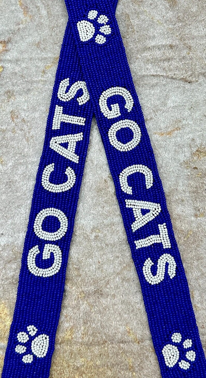 K&ouml;nnte beinhalten: Ein blaues Perlenband mit wei&szlig;en Perlen, die "GO CATS" buchstabieren, und einem Pfotenabdruck-Design.