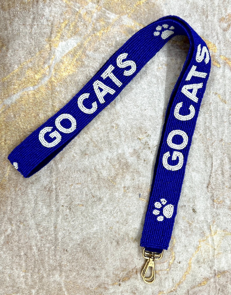 K&ouml;nnte beinhalten: Ein blaues Perlenband mit wei&szlig;en Perlen, die "GO CATS" und Pfotenabdr&uuml;cke buchstabieren. Das Band hat einen goldenen Metallverschluss.