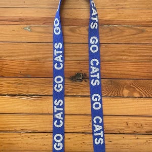 K&ouml;nnte beinhalten: Blau-wei&szlig;er Perlenriemen mit dem Text "GO CATS GO CATS", der auf beiden Seiten wiederholt wird. Der Riemen hat goldfarbene Metallschlie&szlig;en an jedem Ende.