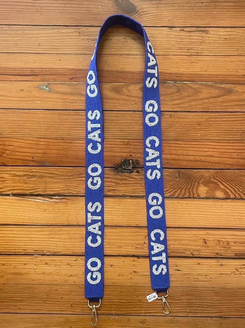 K&ouml;nnte beinhalten: Blau-wei&szlig;er Perlenriemen mit dem Text "GO CATS GO CATS", der auf beiden Seiten wiederholt wird. Der Riemen hat goldfarbene Metallschlie&szlig;en an jedem Ende.