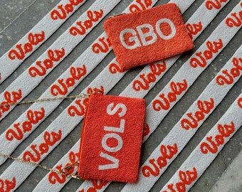 May include: Two orange beaded purses with chain straps. One purse says "VOLS" and the other says "GBO". The purses are on a white background with orange beaded strips that say "Vols" repeated.