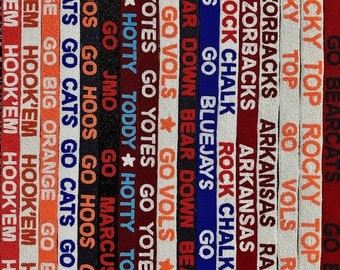 May include: A close-up of a collection of colorful woven fabric bands with various college team names and logos, including "Go Vols", "Go Cats", "Go Hoos", "Razorbacks", "Bluejays", "Bear Down", "Hook'em", "Go Big Orange", "Top Rocky", "Go Yotes", "Go Bears", "Go Todd", "Hotty Toddy", "Rock Chalk", "Arkansas Razorbacks", and "Go Vols".
