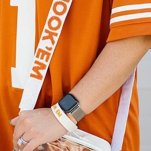 Könnte beinhalten: Orangefarbenes Trikot-Shirt mit einer großen weißen Nummer 1. Ein weißer Umhänge-Taschenriemen mit den Worten "HOOK 'EM" in Orange. Eine transparente Umhängetasche mit weißem Besatz. Eine Smartwatch mit silbernem Armband am Handgelenk.