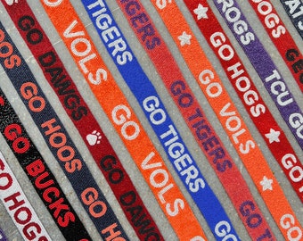 May include: A collection of colorful beaded straps with various college team names and logos, including "Go Vols", "Go Tigers", "Go Dawgs", "Go Bucks", "Go Hoos", and "Go Cats".