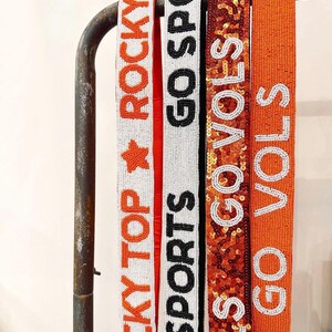 May include: Four beaded belts with different designs. The first belt is white with orange beads and says "Rocky Top Rocky". The second belt is white with black beads and says "Go Sports". The third belt is orange with gold sequins and says "Go Vols". The fourth belt is orange with white beads and says "Go Vols".