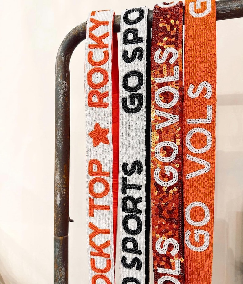 May include: Four beaded belts with different designs. The first belt is white with orange beads and says "Rocky Top Rocky". The second belt is white with black beads and says "Go Sports". The third belt is orange with gold sequins and says "Go Vols". The fourth belt is orange with white beads and says "Go Vols".