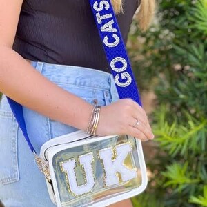 K&ouml;nnte beinhalten: Eine transparente Umh&auml;ngetasche aus Kunststoff mit einem wei&szlig;en Riemen und den Buchstaben "UK" in Wei&szlig; mit goldenen Verzierungen. Der Riemen ist mit den Worten "Go Cats Go" in wei&szlig;en Perlen auf blauem Hintergrund verziert.