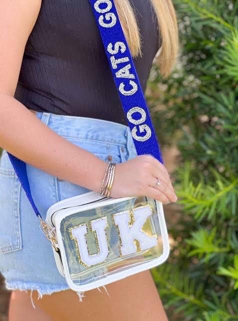 K&ouml;nnte beinhalten: Eine transparente Umh&auml;ngetasche aus Kunststoff mit einem wei&szlig;en Riemen und den Buchstaben "UK" in Wei&szlig; mit goldenen Verzierungen. Der Riemen ist mit den Worten "Go Cats Go" in wei&szlig;en Perlen auf blauem Hintergrund verziert.