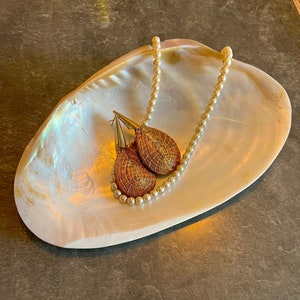 Puede incluir: Un plato en forma de concha blanca nacarada contiene un collar y pendientes dorados. Los pendientes tienen forma de lágrima con un diseño tejido. El collar es una cadena de pequeñas cuentas redondas de color crema. El plato tiene un brillo iridiscente.