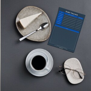 Puede incluir: Vista superior de un rastreador de hábitos azul oscuro con las palabras "Habit Tracker" en blanco. Una porción de pastel está en un plato gris con una cuchara. Una taza de café y un par de gafas también están sobre la mesa.