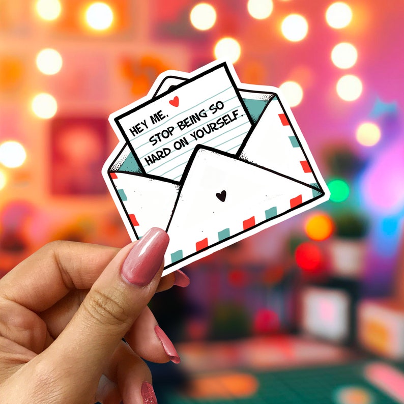 May include: A white sticker in the shape of an envelope with a letter inside. The letter reads "HEY ME. STOP BEING SO HARD ON YOURSELF." The envelope has red and teal accents and a small black heart. The sticker is held by a hand with pink nail polish.