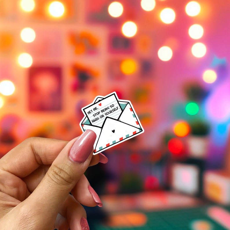 May include: A white sticker in the shape of an envelope with a small heart and red and teal trim. The sticker contains a note that reads "Hey Me. Stop Being So Hard On Yourself."