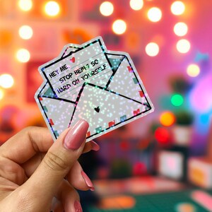 May include: A holographic sticker in the shape of an envelope with a message that reads "Hey Me, Stop Being So Hard On Yourself." The sticker has a heart and a dotted pattern.
