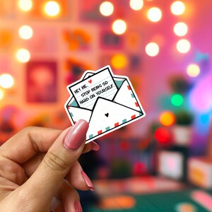 May include: A white sticker in the shape of an envelope with a heart on the front. The envelope contains a note that reads "Hey me. Stop being so hard on yourself." The envelope has red and teal trim.