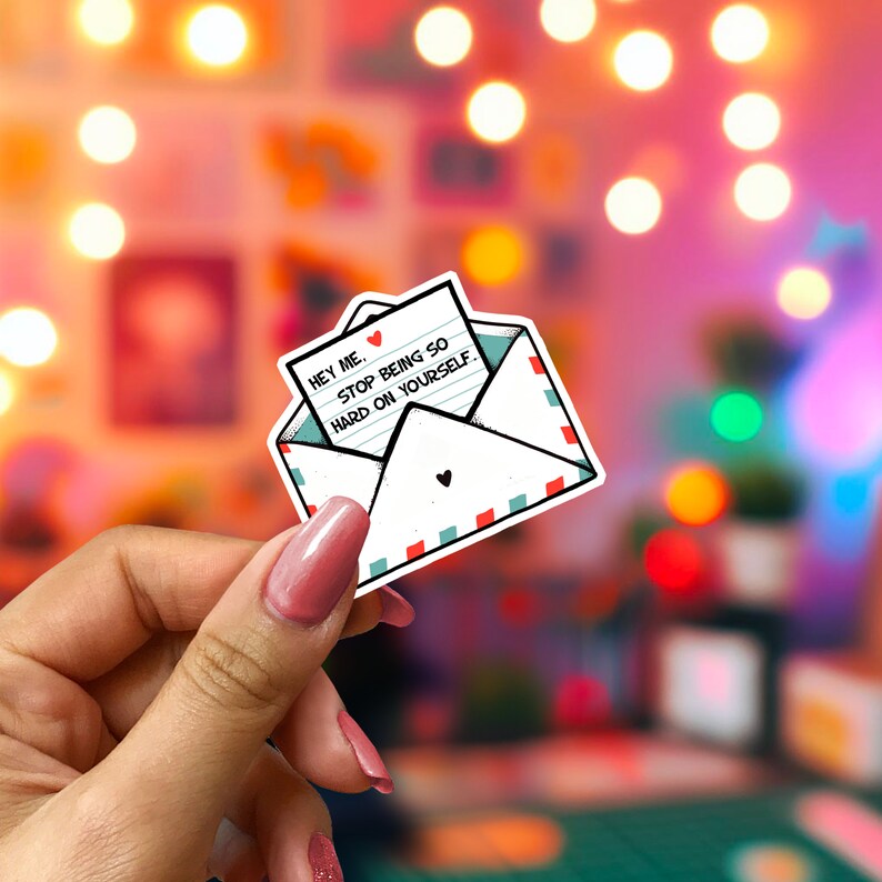 May include: A white sticker in the shape of an envelope with a heart on the front. The envelope contains a note that reads "Hey me. Stop being so hard on yourself." The envelope has red and teal trim.