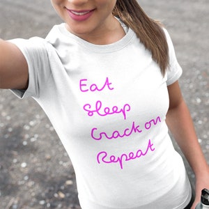 Op de afbeelding: Wit T-shirt met de tekst "Eat Sleep Crack On Repeat" in roze cursief.