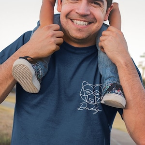 Op de afbeelding: Een man draagt een donkerblauw T-shirt met een wit geometrisch berenontwerp en het woord "Daddy" eronder. Hij draagt een jonge jongen op zijn schouders. De jongen draagt een wit T-shirt en een blauwe jeans.