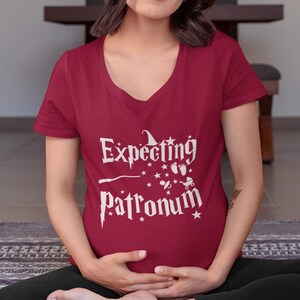 May include: A red v-neck t-shirt with the text "Expecting Patronum" in white. The text is in a stylized font and includes a wand, baby footprints, a baby rattle, and a baby carriage.
