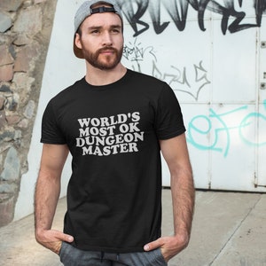 Könnte beinhalten: Ein schwarzes T-Shirt mit weißem Text, der "WORLD'S MOST OK DUNGEON MASTER" lautet.