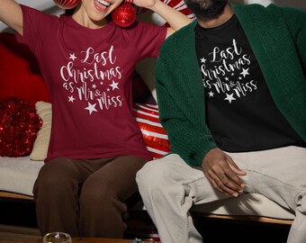 Op de afbeelding: Een stel draagt bijpassende kerst-T-shirts met de tekst "Last Christmas as Mr & Miss" in witte letters. De vrouw draagt een bordeauxrood shirt en de man een zwart shirt. Ze houden kerstballen vast.