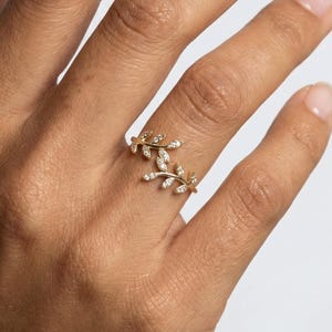 Può includere: Un anello dorato con un design a foglie, ornato da piccole gemme scintillanti. L'anello avvolge il dito, creando un aspetto delicato ed elegante. L'anello è mostrato su una mano con uno sfondo neutro.