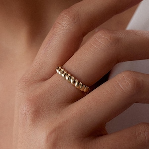May include: A gold ring with a textured, beaded design. The ring is worn on a finger, showcasing its intricate details and warm, metallic colour. The ring's design features a series of small, rounded beads, creating a continuous pattern around the band.