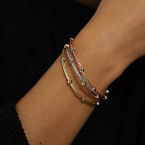 Könnte beinhalten: Drei zarte Armbänder in Gold-, Silber- und Roségoldtönen sind an einem Handgelenk gestapelt. Jedes Armband hat kleine, gleichmäßig verteilte Perlen und ein geknotetes Design. Die Armbänder sind vor einem dunklen Hintergrund platziert.