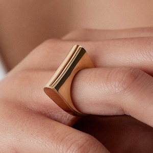 Pode incluir: Um anel dourado com um design retangular. O anel tem uma superfície lisa e polida e é usado no dedo. O design do anel é moderno e minimalista, com linhas limpas e uma aparência elegante. O anel é uma peça de joalharia.