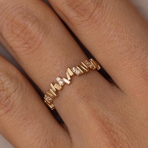 Può includere: Un anello color oro con un design a zig-zag, incastonato con piccole gemme trasparenti. L'anello è mostrato su una mano, evidenziando il suo motivo intricato e la sua brillantezza. Il design dell'anello è moderno ed elegante.