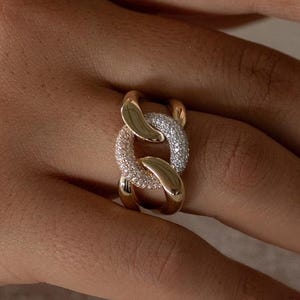 Può includere: Un anello dorato con un design a catena. Un anello è incastonato con piccole pietre preziose trasparenti, creando un contrasto scintillante con l'oro lucido. L'anello è indossato su una mano con unghie curate.