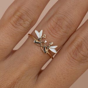 Pode incluir: Anel dourado com um design delicado de duas figuras entrelaçadas com asas de esmalte branco. O anel é mostrado em uma mão, em um fundo rosa claro.