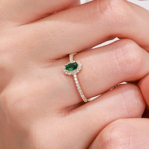 Könnte beinhalten: Ein goldener Ring mit einem grünen ovalen Edelstein, der von kleineren Diamanten umgeben ist. Der Ring befindet sich an einem Finger.