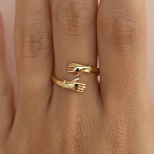 Könnte beinhalten: Ein goldfarbener Ring mit zwei Händen, die sich umarmen. Das Design des Rings umschließt den Finger, wobei die Hände so positioniert sind, als würden sie sich gegenseitig halten. Der Ring besteht aus glänzendem, reflektierendem Metall.
