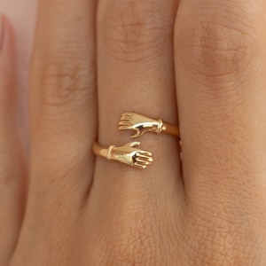 Può includere: Un anello color oro con un design di due mani che si abbracciano. La fascia dell'anello si curva attorno al dito, con ogni mano dettagliata con le dita e un polsino. L'anello è realizzato in metallo lucido e lucido.