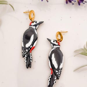 May include: A pair of handcrafted earrings featuring a woodpecker design. The earrings are black and white with red accents, and gold-colored earring posts. The earrings are set against a white background with sprigs of lavender.