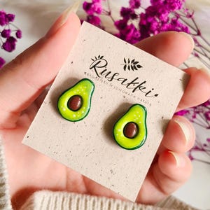 Puede incluir: Pendientes con forma de aguacate sobre una tarjeta beige. Los pendientes son verdes con una semilla marrón y motas blancas. La tarjeta tiene el texto "Rusatki" y un diseño floral. Los pendientes son sostenidos por una persona.